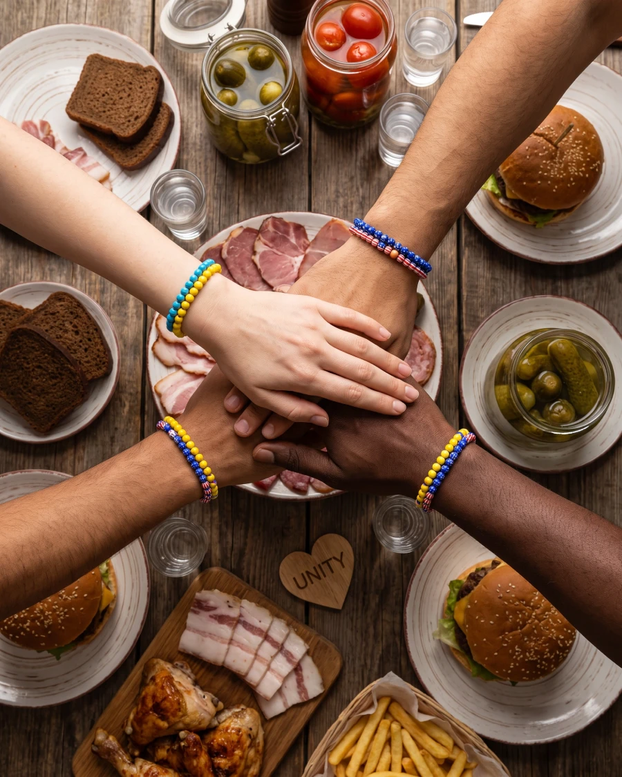 Overhead view of a multicultural dinner table with Slavic zakuski, American comfort foods, vodka glasses, and diverse hands stacked together.