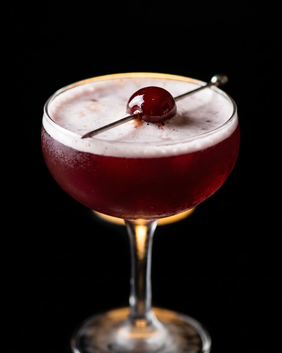 A cherry sour in a coupe glass — vibrant deep crimson-red liquid with a foam cap and brandied cherry