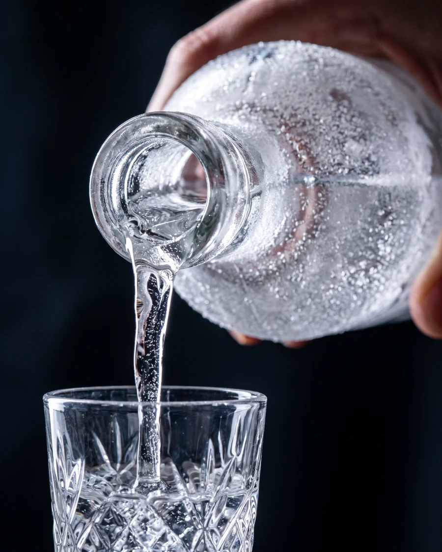A frosted vodka bottle being poured into a small crystal shot glass — the liquid nearly frozen, viscous and clear